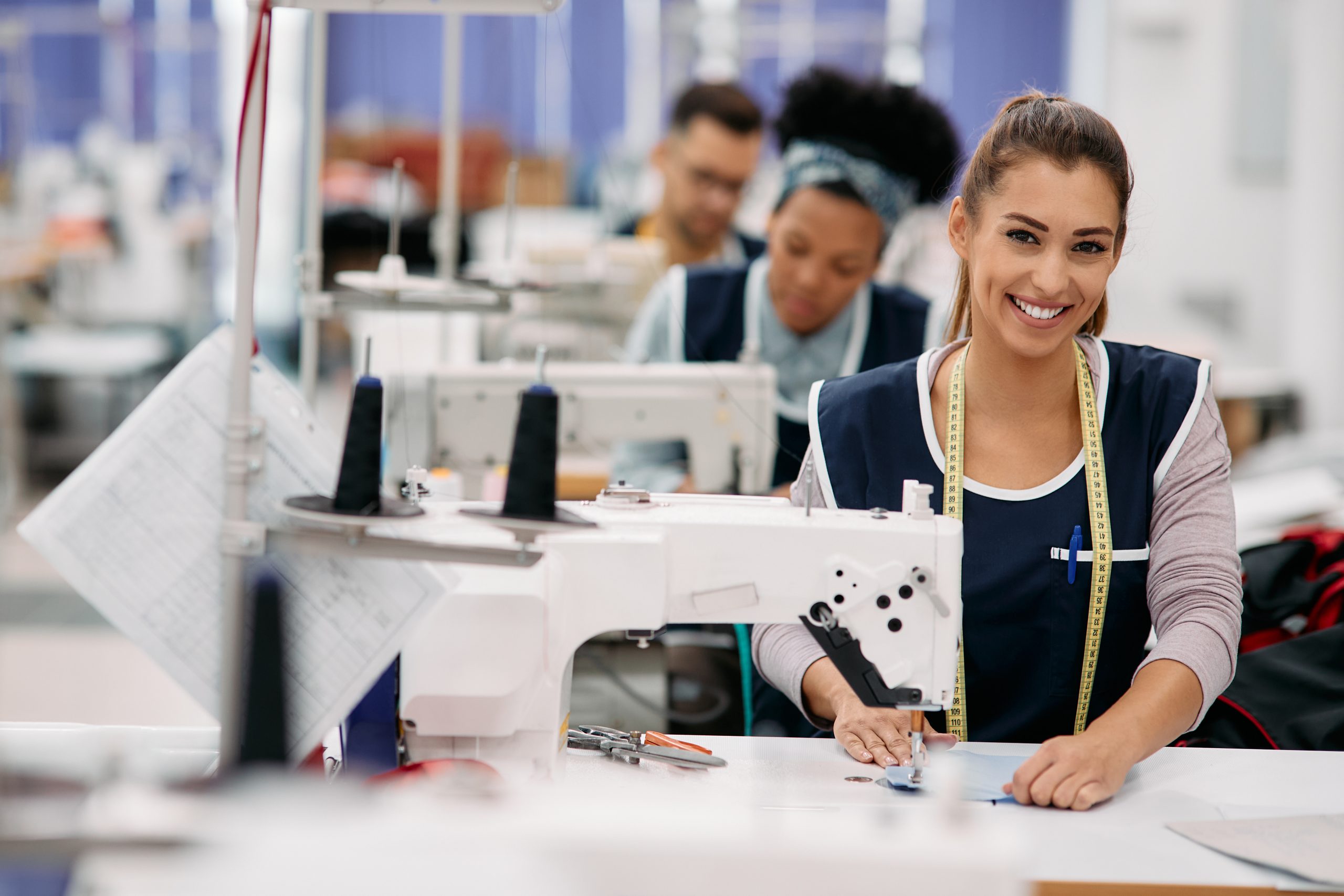 Young happy seamstress works at production line in factory and looks at camera.